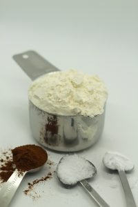 silver measuring cup filled with flour and three separate silver measuring spoons filled with cinnamon salt and baking soda on white surface