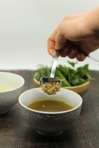 hand holding a heart shaped measuring spoon revealing garlic above olive oil white dish plus herbs and lemon juice in two separate white bowls in background on dark wooden surface