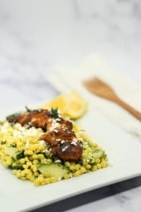 Yellow couscous salad with cucumber and herbs topped with sliced blackened chicken and feta cheese on a white plate with lemon wedges and wooden fork