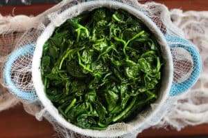 Blanched spinach over a cheesecloth in a white bowl with light blue handles on brown surface