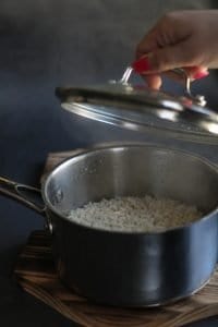 Bibimbap - silver pot filled with steamed rice on wooden base on grey surface and hand with pink nails lifting the pot lid