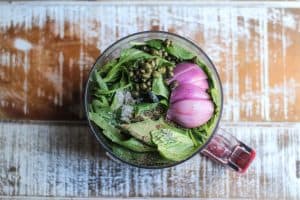 Basil vinaigrette ingredients in a food processor on a white and brown wooden surface.