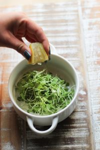 hand squeezing lemon juice onto micro arugula salad in a while bowl on brown and white wooden surface