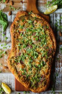 cheesy flatbread pizza with salami and honey goat cheese almond pesto topped with micro arugula salad on a brown cutting board on brown and white wooden surface