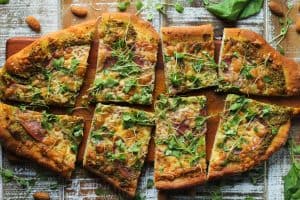 sliced cheesy flatbread pizza with salami and honey goat cheese almond pesto topped with micro arugula salad on a brown cutting board on brown and white wooden surface