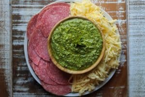 honey goat cheese almond pesto in a wooden bowl in center of plate surrounded by salami slices, shredded cheddar and provolone cheese