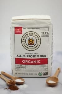 bag of organic King Arthur all-purpose flour next to wooden spoons filled with salt, baking soda, baking powder, and cinnamon on a white marble surface