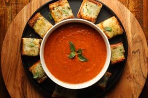 roasted tomato basil and red pepper soup in a white bowl with slices of cheesy garlic bread on a black plate on a brown surface