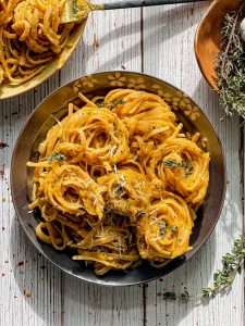 Pasta with Roasted Garlic Butternut Squash Sauce in a gray bowl on a white wooden surface with pasta twisted around fork and thyme sage and garlic bulb in a bowl