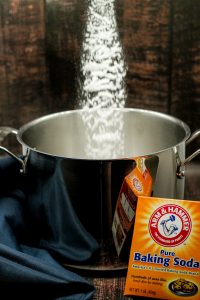 Orange container of Arm & Hammer baking soda being added to a silver stainless steel pot filled with water on a brown wooden surface with a blue napkin on the side
