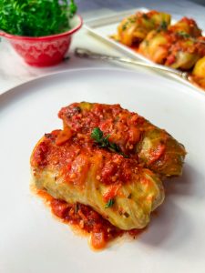 two stuffed cabbage rolls with tomato sauce on a white plate, with a white serving plate with stuffed cabbage rolls and a red bowl filled with parsley in the background