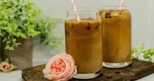 Glass of 4 Ingredient Chaga Mushroom Iced Coffee with Espresso on a dark wooden cutting board with a few whole coffee beans, a white background, and small pink peonie, and a green plant in the background