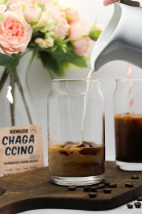 Milk being poured into a glass of 4 Ingredient Chaga Mushroom Iced Coffee with Espresso on a dark wooden cutting board with several scattered whole coffee beans, a white background, a glass vase with a bouquet of pink peonies, and a pink packet of Chagaccino in the background