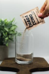 Chagaccino superfood mushroom coffee boost being poured into a glass of 4 Ingredient Chaga Mushroom Iced Coffee with Espresso on a dark wooden cutting board, a white background, and a green plant in the background