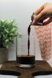 Espresso being poured into a glass of 4 Ingredient Chaga Mushroom Iced Coffee with Espresso on a dark wooden cutting board, a white background, and a green plant & a cup of pink and white striped straws in the background