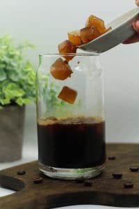 Coffee ice cubes being dumped into a glass of 4 Ingredient Chaga Mushroom Iced Coffee with Espresso, on a dark wooden cutting board, several scattered coffee beans, a white background, and a green plant in the background