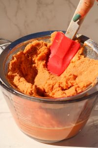 blended watermelon and ginger being strained into a big glass bowl through a fine mesh strainer with a red rubber platula