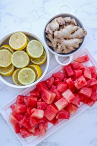 Diced watermelon, halved lemons, and fresh ginger on and in white plates and bowls on a white granite surface