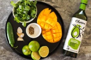 Ingredients for mango sauce on a black plate: Jalapeno, garlic, halved limes, cilantro, mangoes, salt, pepper, chosen foods avocado oil