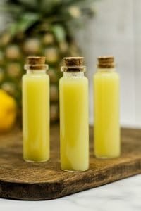 Pineapple Coconut Lemon Ginger Juice Wellness Shots Recipe: three shots in tiny glass vials with cork top on a brown wooden cutting board with a pineapple and lemon blurred in the background