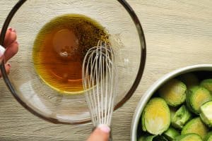 maple syrup being whisked with lemon juice, salt, and pepper and a bowl of halved brussel sprouts on the side.