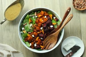 Roasted beetroot and pumpkin salad in a white bowl with mustard vinaigrette, goat cheese, and chopped pecans on the side.