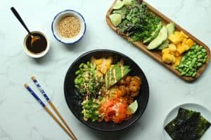 Assembled shrimp poke bowl recipe with sweet soy glaze in a white jar, sesame seeds, and toppings on a plate.