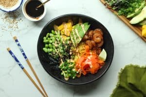 Assembled shrimp poke bowl recipe with sweet soy glaze in a white jar, sesame seeds, and toppings on a plate.