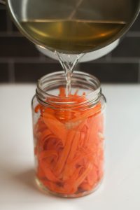 Pickling liquid being poured into a mason jar filled with shaved carrot strips and garlic.