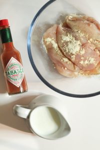 Chicken tenderloins in a bowl with seasoning and a bottle of tobasco and some buttermilk on the side.
