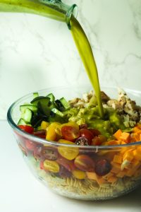 Protein pasta salad with chicken ingredients in glass bowl with lemon parsley vinaigrette being poured on it.