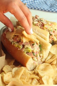 A hand with white nail polish placing salt and vinegar chips on chopped Italian sub sandwiches and a side of chips.