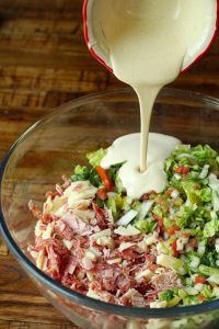 Chopped Italian meats and cheeses on one side of the bowl and chopped vegetables on other. Deli sauce being poured in.