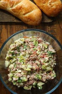 A clear glass bowl with chop Italian sub sandwiches filling and two loaves of sourdough bread on a wooden cutting board.