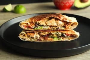 Chicken crunch wrap cut in half and stacked on top of the other half on a black plate, with a cut lime and avocado half in the background.