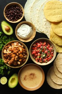 Chicken brunch wrap ingredients scattered in individual bowls, plus tortillas, avocado, cilantro, and a halved lime on a black surface.
