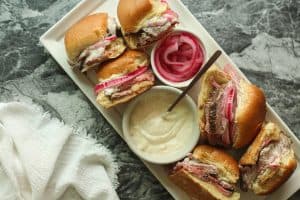 Roast beef sliders on a white rectangular plate with a bowl of garlic horseradish aioli and a bowl of pink colored pickled onions.