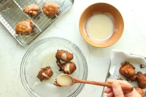 Maple glaze being drizzled on applesauce fritters in a bowl before being tossed and a bowl of glaze on the side.