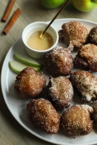 Close up of glazed and powdered sugar dusted applesauce fritters with a bowl of glaze, and cinnamon sticks and whole apples in the back.