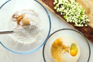 Two separate clear bowls with wet and dry applesauce fritters ingredients and a wooden cutting board with diced apples.