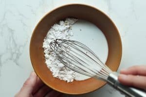 Powdered sugar, maple syrup, and milk being whisked in a golden-brown bowl on a white surface.