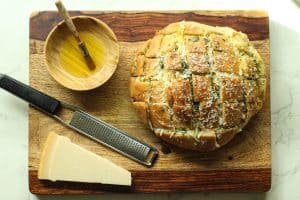 A loaf of stuffed garlic bread with cheese on a cutting board with a bowl of olive oil, a grater, and a block of parmesan.