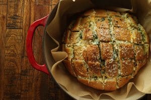 A loaf of stuffed garlic bread topped with parmesan in a parchment paper lined Dutch oven with a red handle on wooden surface.