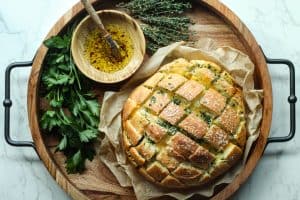 Stuffed garlic bread on a piece of parchment paper next to a bowl wooden bowl with oil and fresh green herbs.