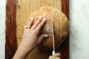 A handholding a round loaf of bread down while the other hand uses a knife to cut lines down the bread on a wooden board.