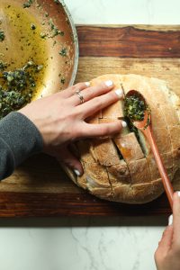 Hands pulling back pieces of the cut bread and spooning garlic butter mixture into the cracks.