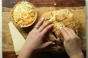 Two hands with white nails stuffing stuffed garlic bread with shredded cheese on a cutting board with a block of parmesan.
