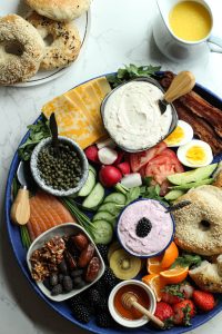 Bagel board charcuterie style with sweet and savory spreads, fruit, vegetables, salmon lox, bacon, eggs, and a pitcher of orange juice.