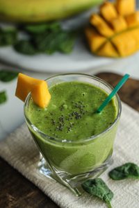 Green protein smoothie sprinkled with chia seeds in a clear glass cup with a cut piece of mango on the rim.