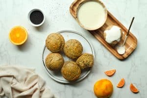 4 whole and one halved orange poppyseed muffins in the center on a white plate with surrounding ingredients and oranges.
